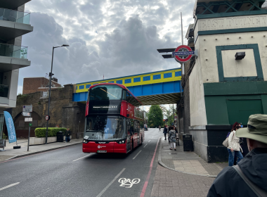 Red double-decker bus on a city street under a blue-and-yellow bridge, with a London Underground roundel sign nearby and pedestrians on the sidewalk.