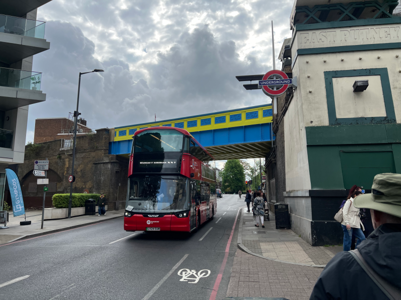 Red double-decker bus on a city street under a blue-and-yellow bridge, with a London Underground roundel sign nearby and pedestrians on the sidewalk.