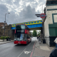 Red double-decker bus on a city street under a blue-and-yellow bridge, with a London Underground roundel sign nearby and pedestrians on the sidewalk.