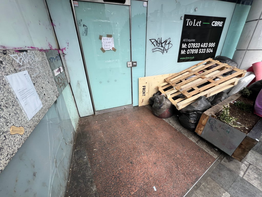 Teal storefront with glass doors, a large CBRE 'To Let' sign, graffiti, and a pile of pallets and black bags outside.