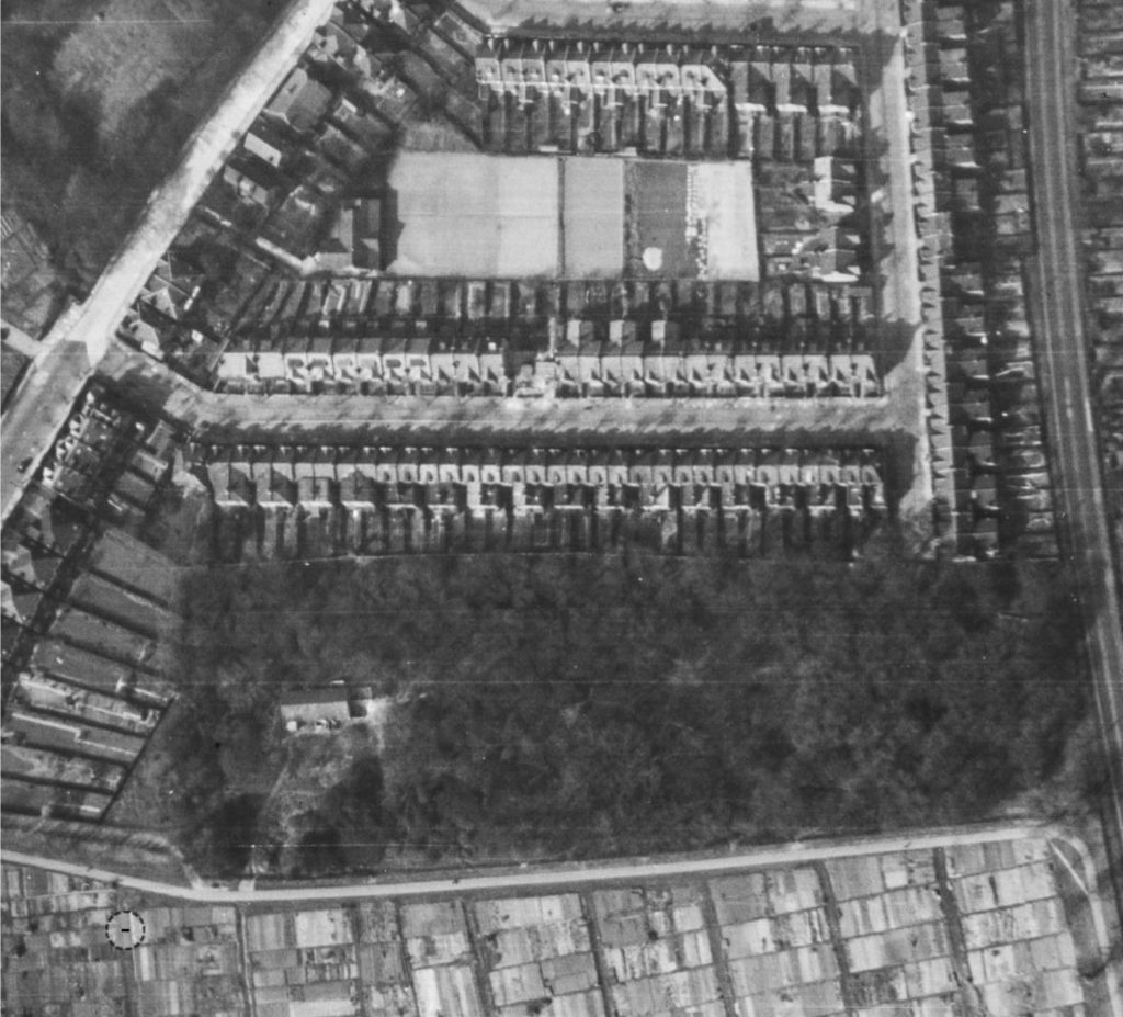 Aerial black-and-white view of dense urban housing with rows of terraced houses surrounding a large rectangular central building and surrounding roads.