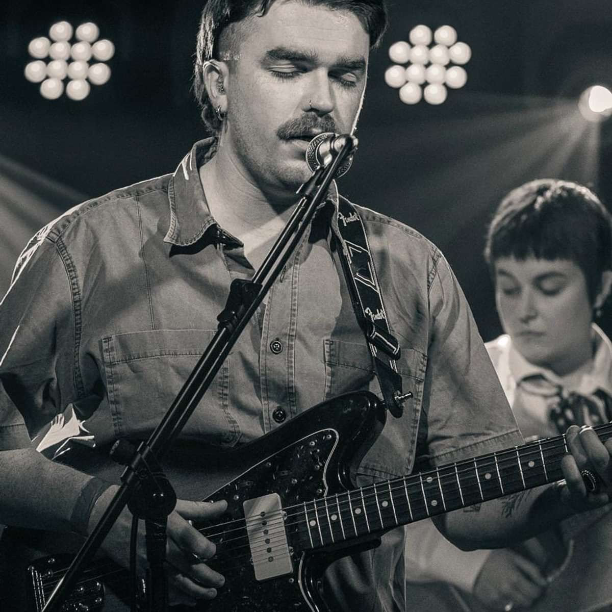 Male musician singing into a microphone while playing a black electric guitar on stage under bright stage lights.