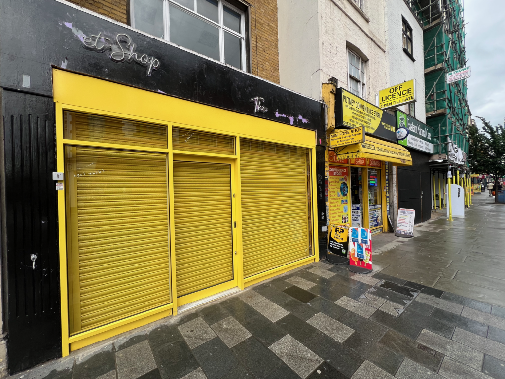 Bright yellow shuttered storefront on an urban street, with black awning and chalky purple signage above.