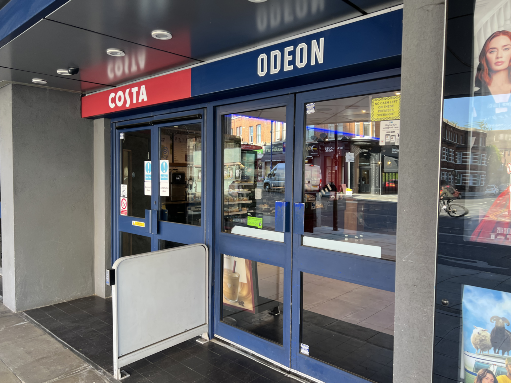 Costa and Odeon storefront with blue doors and red Costa sign; glass entrance reflecting street scenes outside.