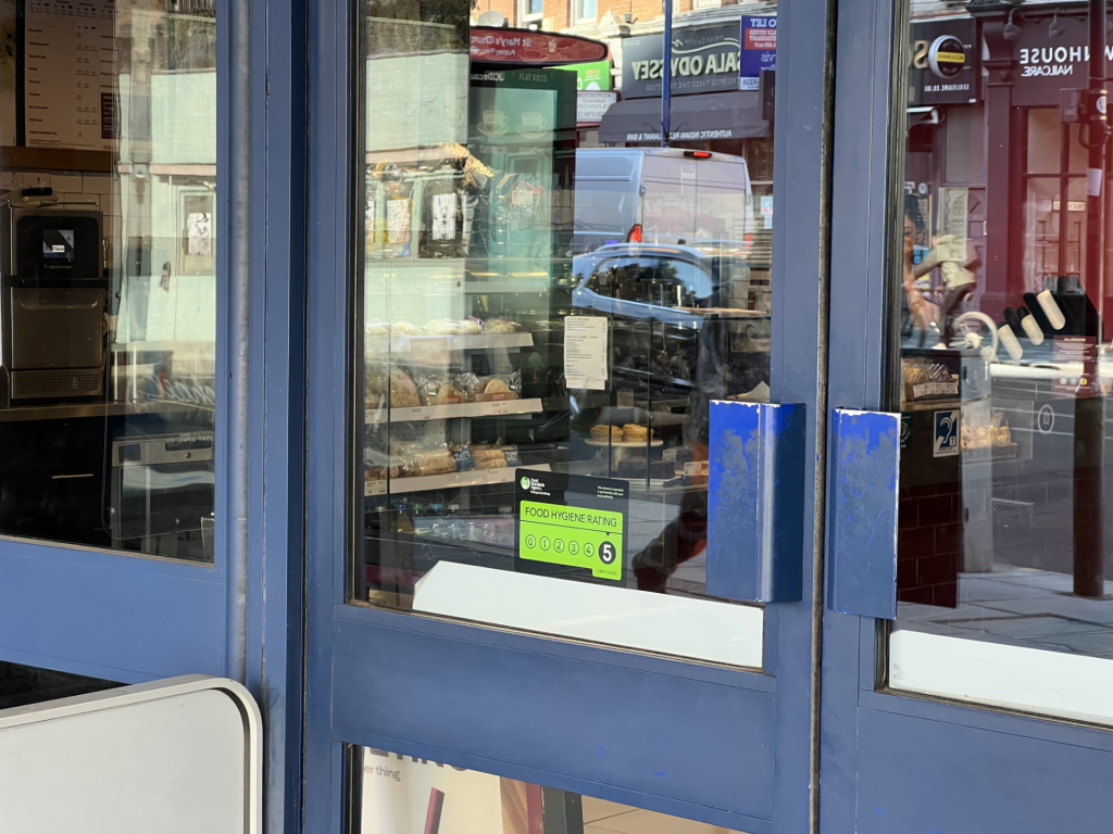 Blue-framed storefront window displaying pastries inside, with a green food hygiene rating sticker visible inside the glass and street reflections outside