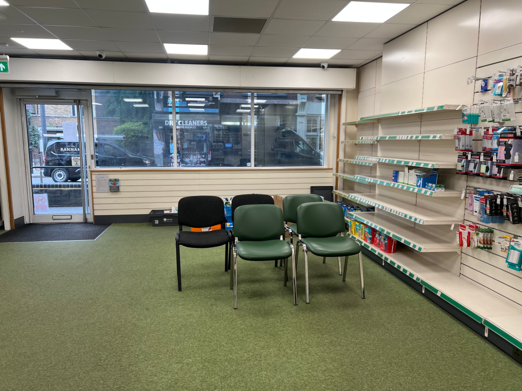 Pharmacy or clinic seating area with four green and black chairs in the center and shelves of products along the right wall; glass storefront and door to street on the left.