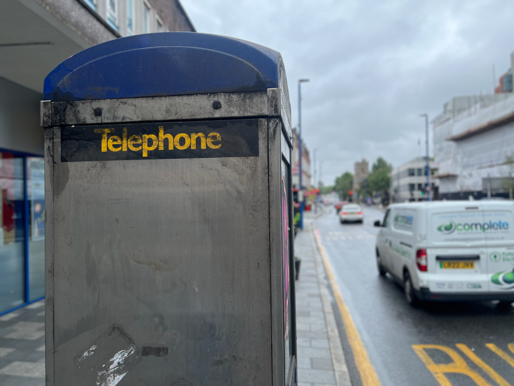 Close-up of a weathered public telephone booth with a blue curved top and a black panel reading 'Telephone' in yellow, on a city sidewalk.