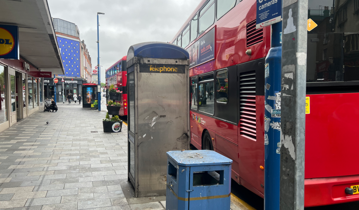 Red double-decker bus parked along a city sidewalk with a blue public phone booth and storefronts on the left side of the street.