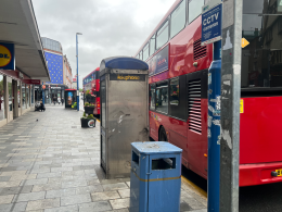 Red double-decker bus parked along a city sidewalk with a blue public phone booth and storefronts on the left side of the street.