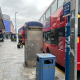 Red double-decker bus parked along a city sidewalk with a blue public phone booth and storefronts on the left side of the street.
