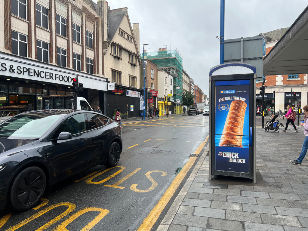 City street with shopfronts (M&S Food) on the left, pedestrians and a parked car in the foreground, and a bus shelter with a large chicken advertisement on the right.