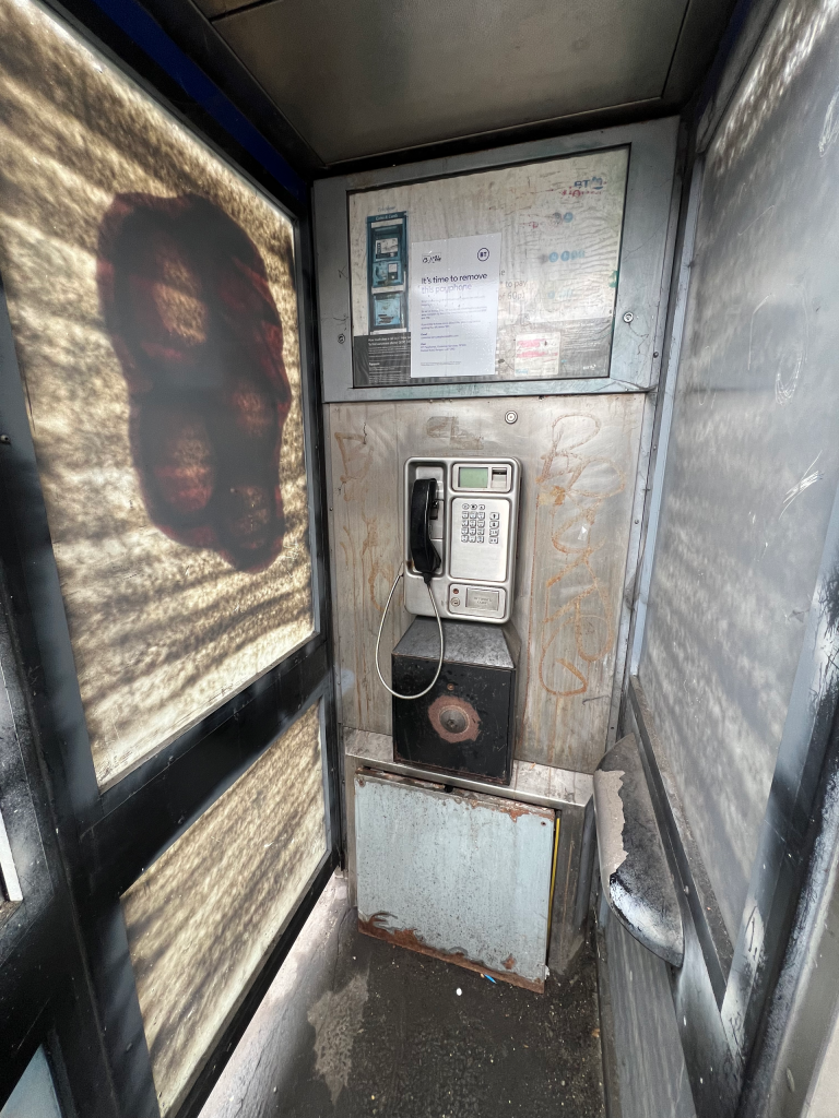 Old public payphone in a narrow, graffiti-streaked booth with a corded handset and keypad on a dirty metal panel back wall.