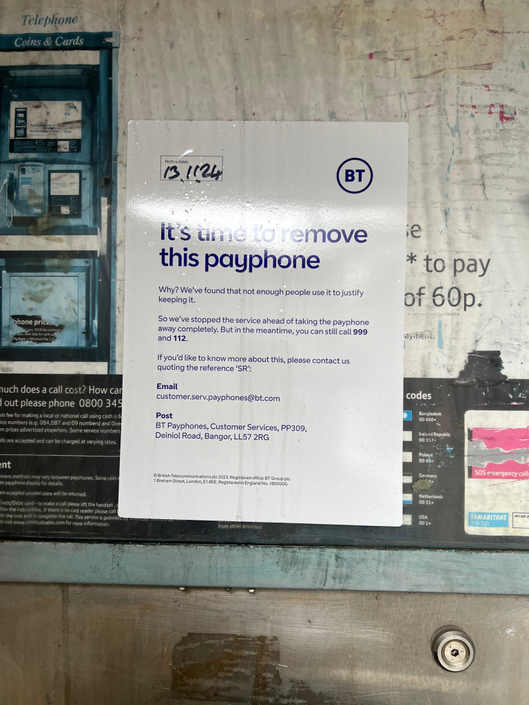 White BT notice on a weathered wall announcing the payphone removal, with a blue phone booth framing on the left side of the image.
