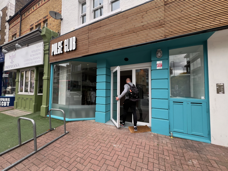 Man with backpack entering Pulse Club, a turquoise storefront on a city street.