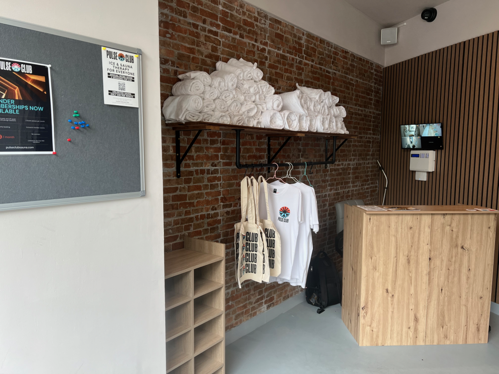 Brick‑wall interior with a long shelf holding neatly rolled white towels above clothing with Pulse Club logos, wooden reception desk nearby.