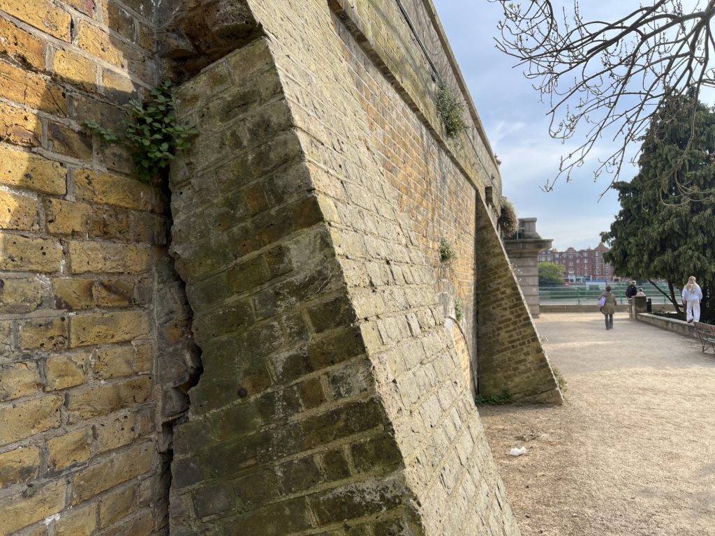 Angled, mossy stone wall with small plants growing in the gaps, beside a riverside promenade and distant pedestrians.