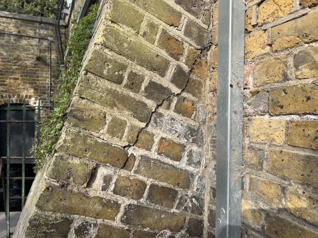 Slanted mossy brick wall with a vertical metal post and ivy along the left edge in an outdoor setting.