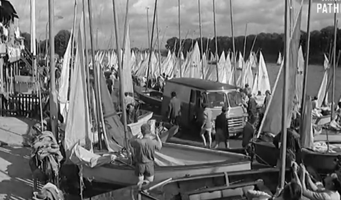 Busy marina with numerous sailboats and tall masts; people walk along the dock near a van.