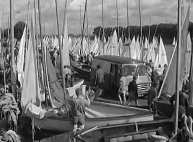 Busy marina with numerous sailboats and tall masts; people walk along the dock near a van.