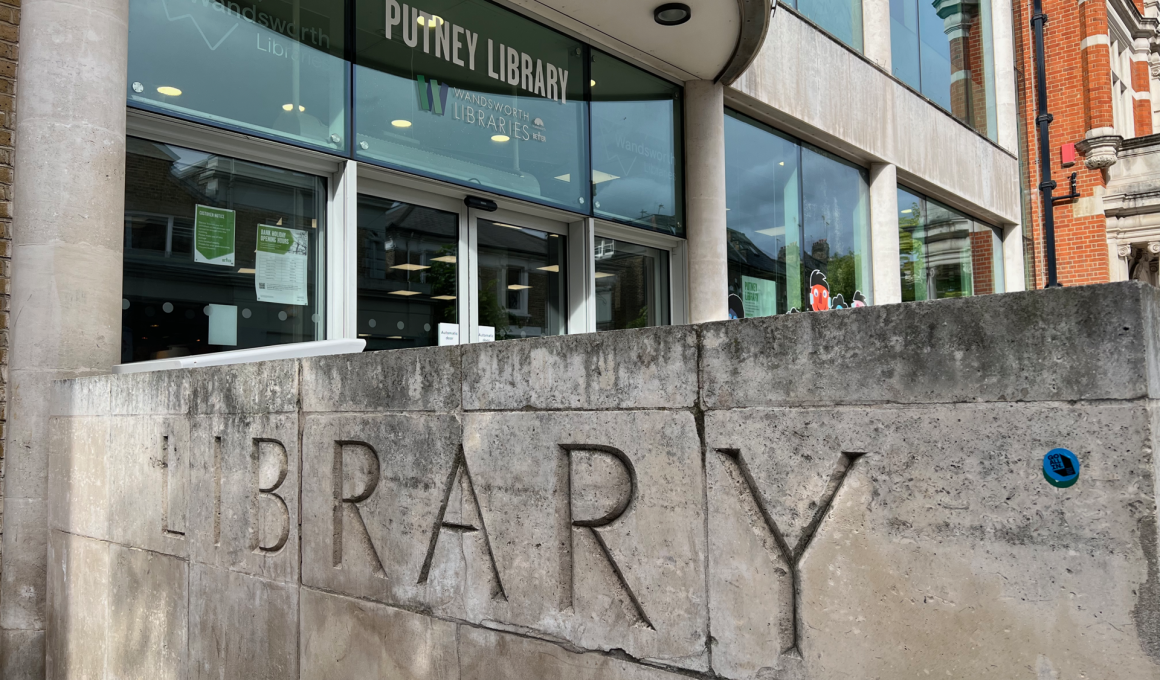 Facade of Putney Library showing a curved glass entrance, large 'PUTNEY LIBRARY' sign, and a textured stone wall with big carved 'LIBRARY' letters.