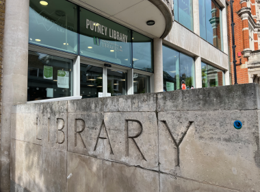 Facade of Putney Library showing a curved glass entrance, large 'PUTNEY LIBRARY' sign, and a textured stone wall with big carved 'LIBRARY' letters.