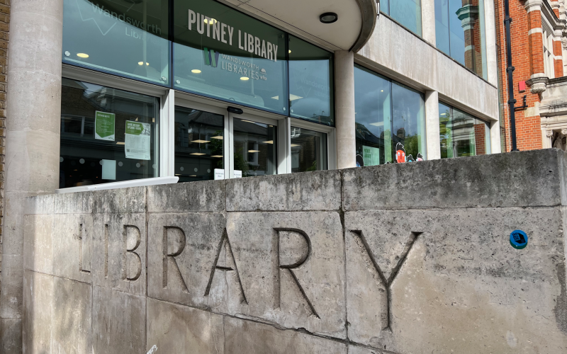 Facade of Putney Library showing a curved glass entrance, large 'PUTNEY LIBRARY' sign, and a textured stone wall with big carved 'LIBRARY' letters.