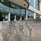 Facade of Putney Library showing a curved glass entrance, large 'PUTNEY LIBRARY' sign, and a textured stone wall with big carved 'LIBRARY' letters.
