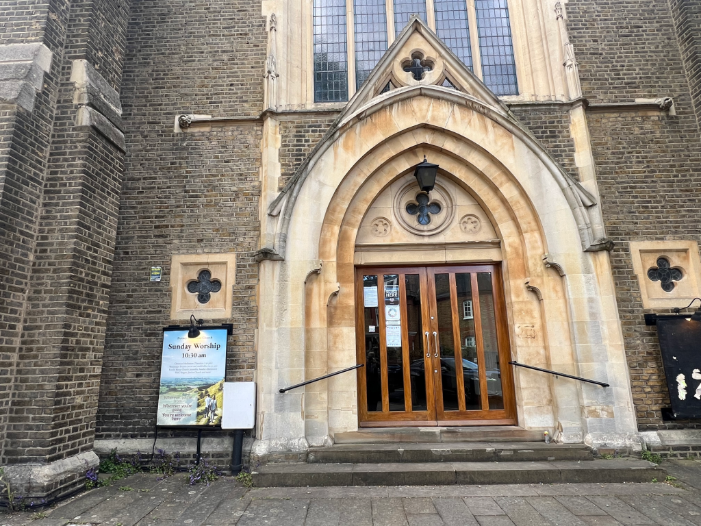 Stone church facade featuring a pointed arch doorway with wooden double doors, flanked by brick walls and stained-glass windows.