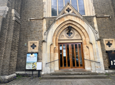 Stone church facade featuring a pointed arch doorway with wooden double doors, flanked by brick walls and stained-glass windows.