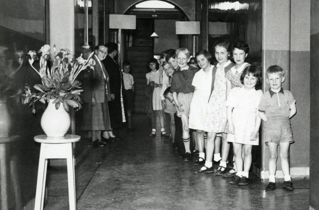Line of smiling children standing in a hallway in a single file, with adults at left near the wall and a potted plant nearby.