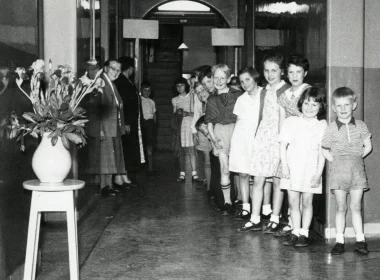 Line of smiling children standing in a hallway in a single file, with adults at left near the wall and a potted plant nearby.