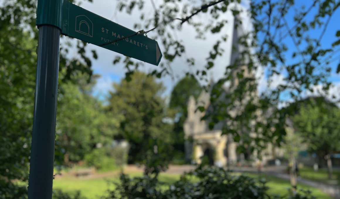 Green signpost pointing to St Margaret's, Putney, mounted on a teal metal pole with leafy background.