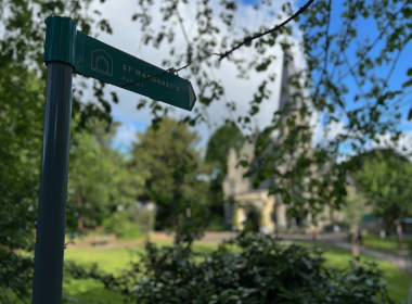 Green signpost pointing to St Margaret's, Putney, mounted on a teal metal pole with leafy background.