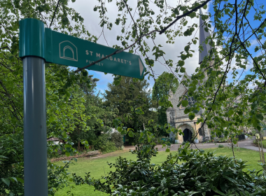 Green directional sign on a blue post, pointing toward St Margaret's, with a church spire visible through leafy branches in a park setting.