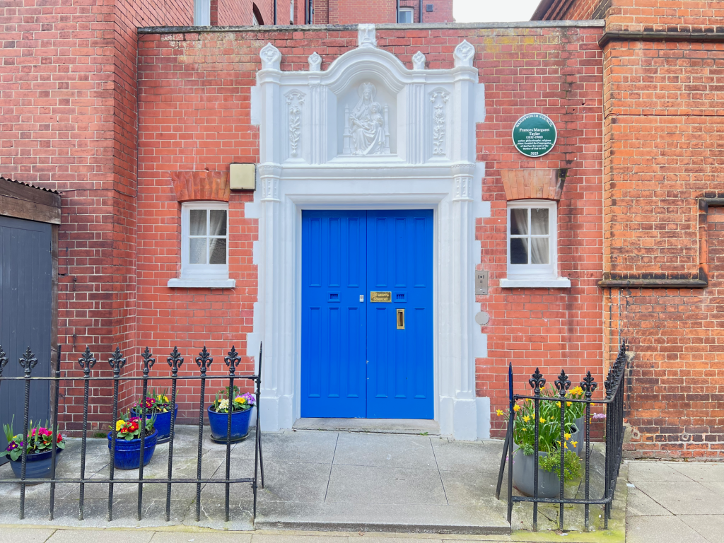 Bright blue door framed by a white ornate doorway in a red brick townhouse, with flower pots along a black iron fence outside.