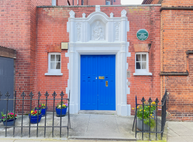 Bright blue door framed by a white ornate doorway in a red brick townhouse, with flower pots along a black iron fence outside.