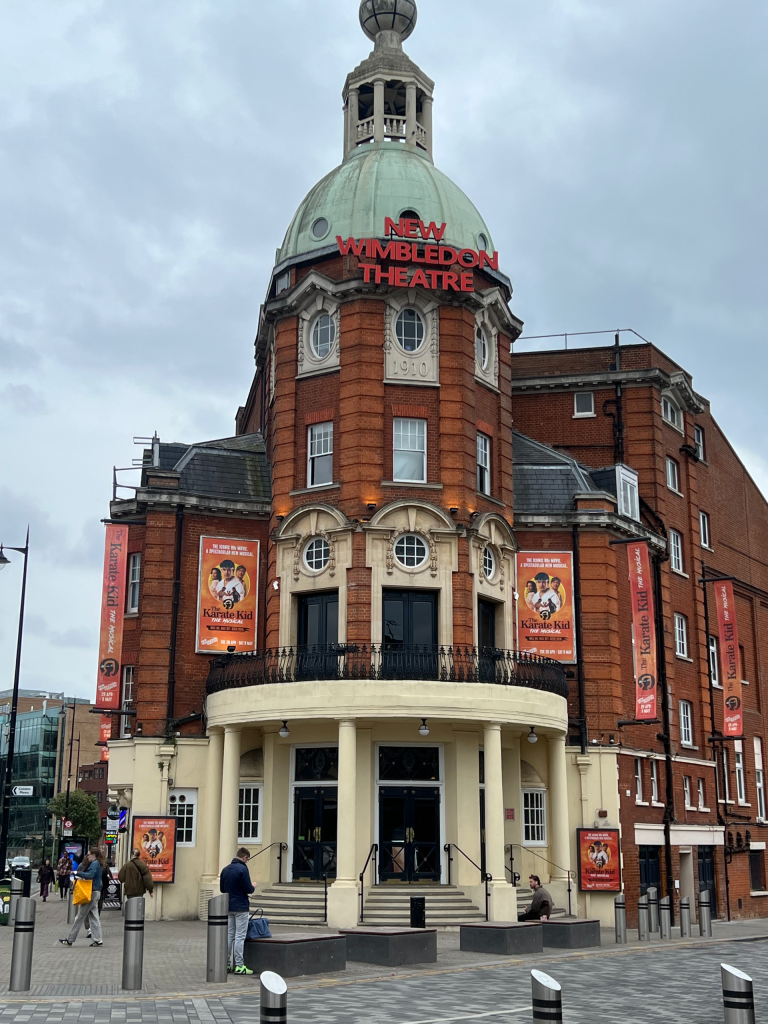The New Wimbledon Theatre: red-brick building with a green domed top and red signage, banners promoting The Karate Kid musical along the front.