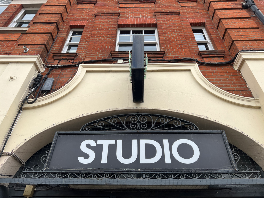 Brick building with red brick facade, cream archway, and a large black sign reading STUDIO under a metal beam and decorative ironwork.