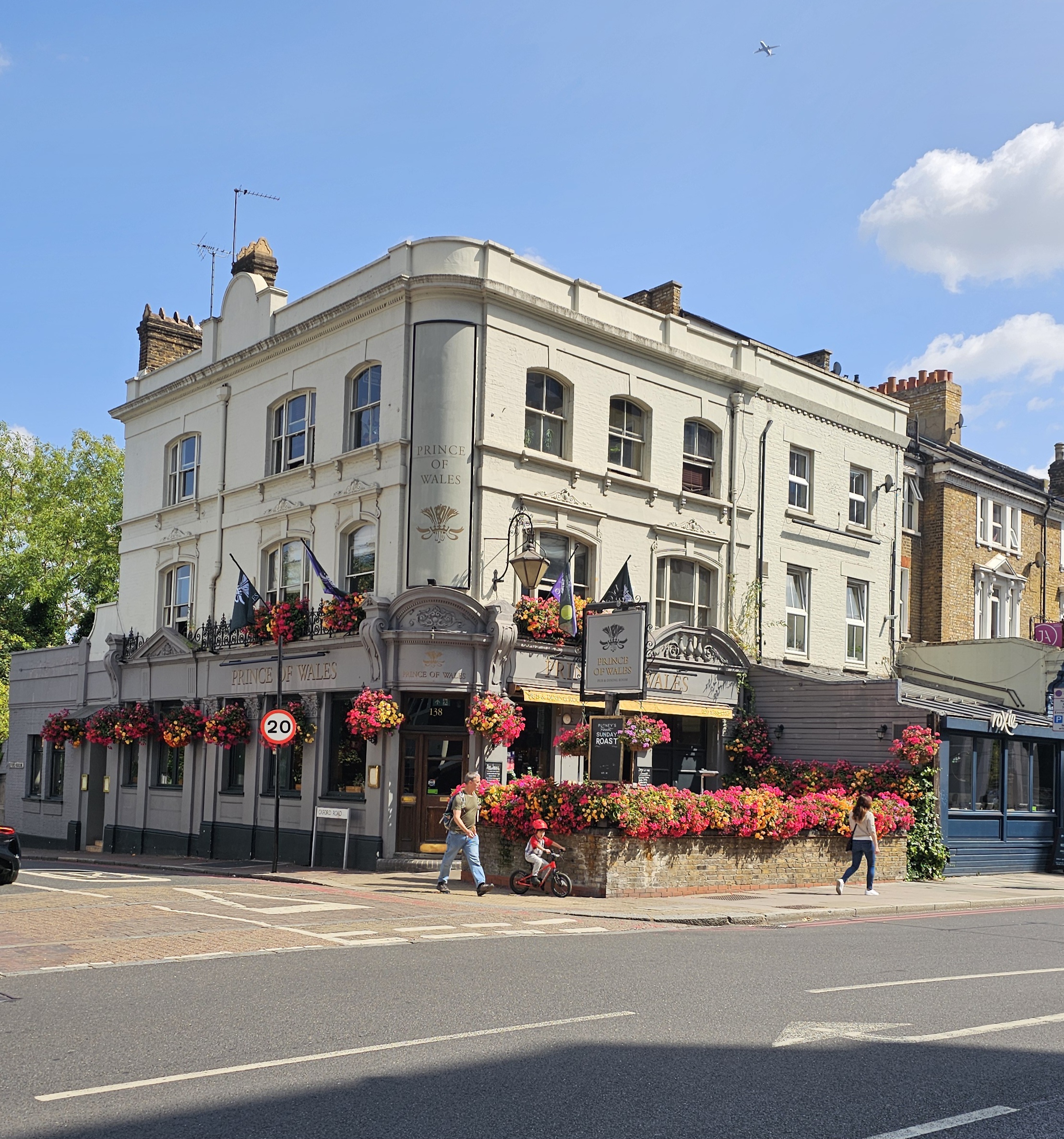 White, four-story pub building named 'Prince of Wales' with ornate facade and hanging flower baskets outside on a sunny street corner (informative).