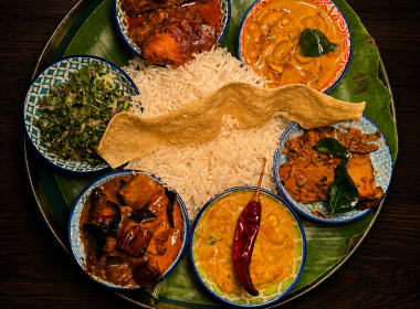 Indian thali on a banana leaf tray: white rice in the center surrounded by several bowls of curry, greens, and a papadam.