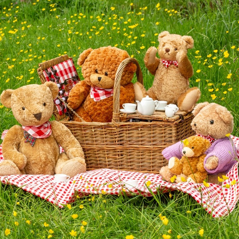 Teddy bears having a picnic on a red-and-white checkered blanket with a wicker basket and a tea set in a sunny meadow.