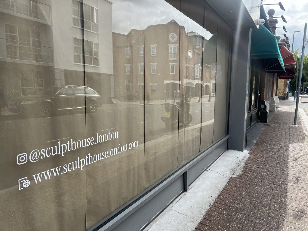 Shopfront window with reflective glass showing white text for social media and a URL (sculptphouse.london) on a city street sidewalk.
