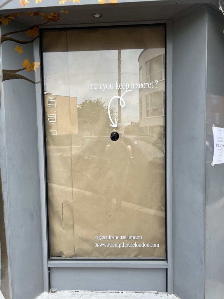 storefront window with grey frame and autumn leaves; white text reads 'can you keep a secret?' with a curved arrow pointing to a small dark circular hole; bottom shows social handles '@sculpthouse.london' and 'www.sculpthouselondon.com'
