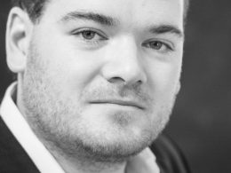 Close-up black-and-white portrait of a man with light stubble looking at the camera, wearing a collared shirt and dark jacket.