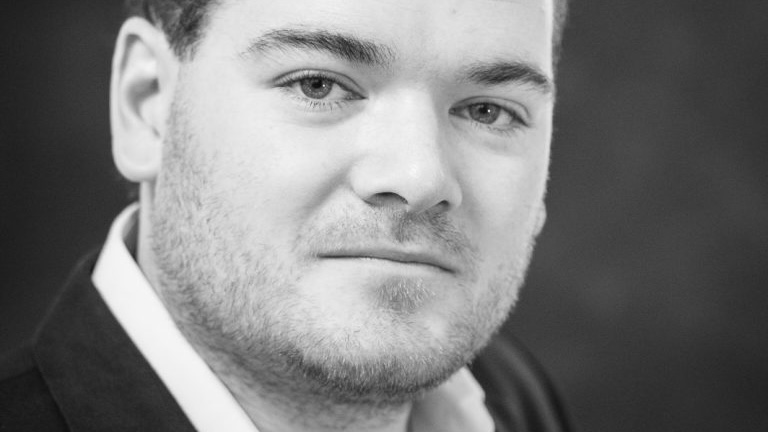 Close-up black-and-white portrait of a man with light stubble looking at the camera, wearing a collared shirt and dark jacket.