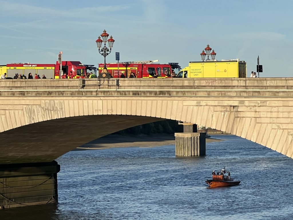 Emergency vehicles on a city bridge: red rescue engines and a yellow incident-response unit, with people walking along the railing and a small boat in the water nearby.