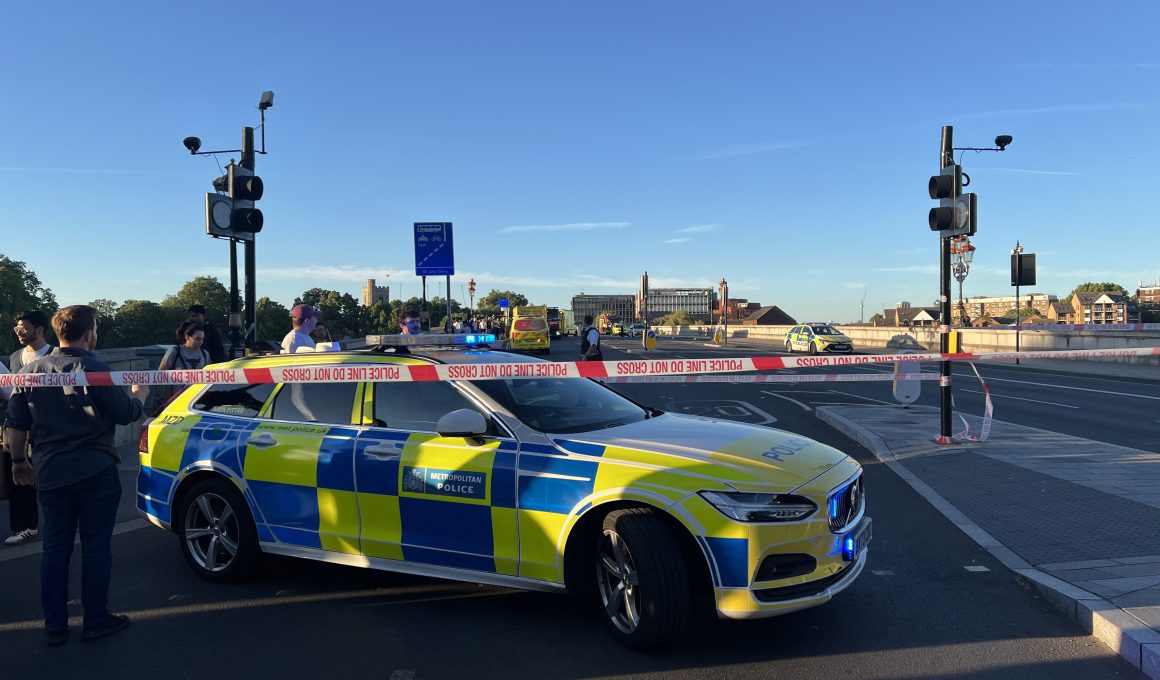 Police car with blue and yellow chequered livery behind police tape reading 'POLICE LINE DO NOT CROSS' on a city street