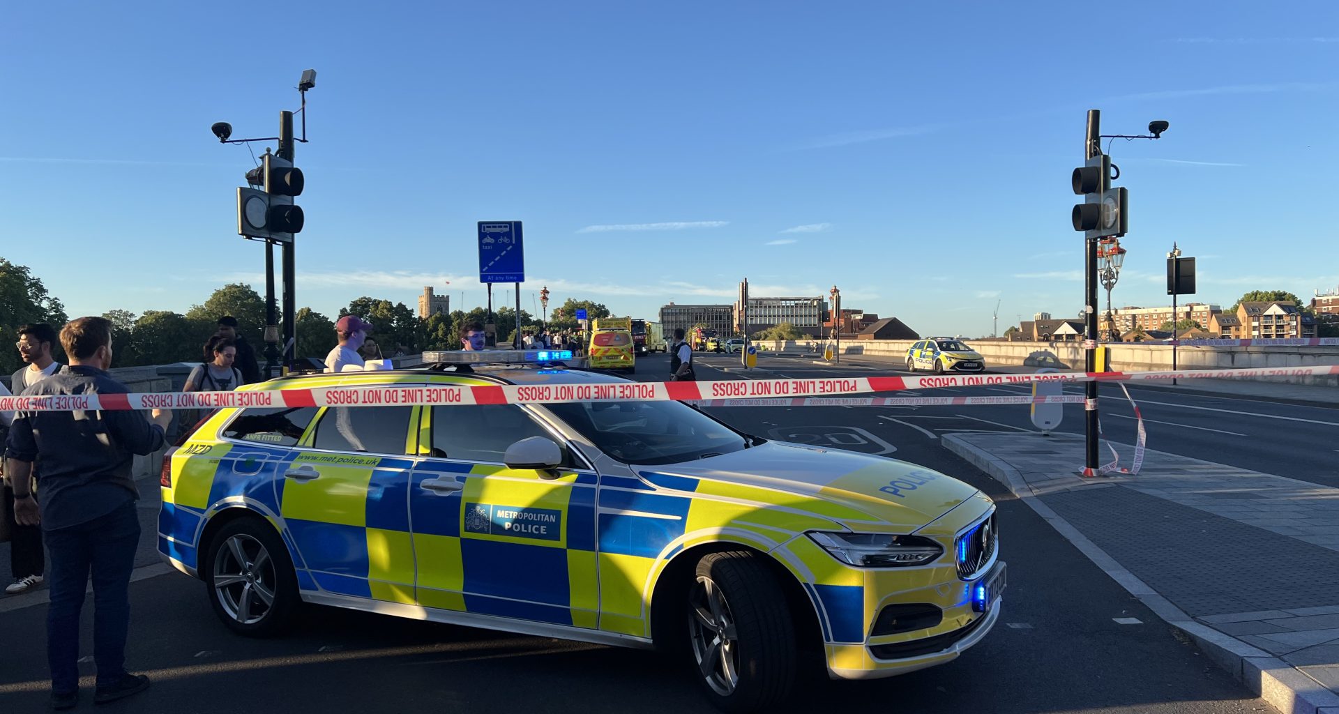Police car with blue and yellow chequered livery behind police tape reading 'POLICE LINE DO NOT CROSS' on a city street