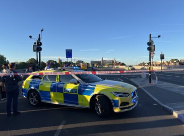Police car with blue and yellow chequered livery behind police tape reading 'POLICE LINE DO NOT CROSS' on a city street
