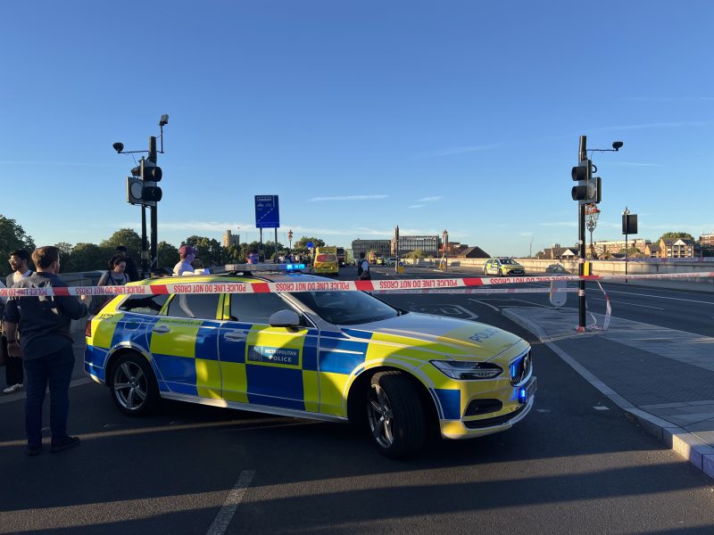 Police car with blue and yellow chequered livery behind police tape reading 'POLICE LINE DO NOT CROSS' on a city street